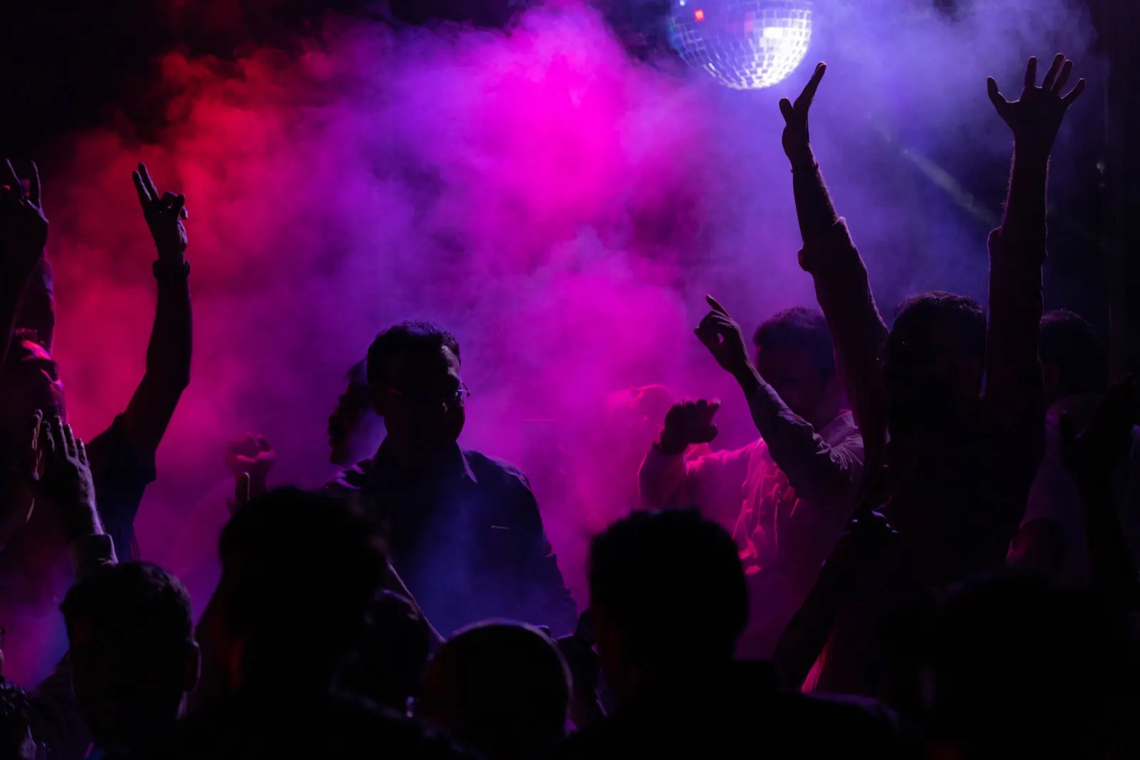 Concert crowd with raised hands under a disco ball in pink smoke