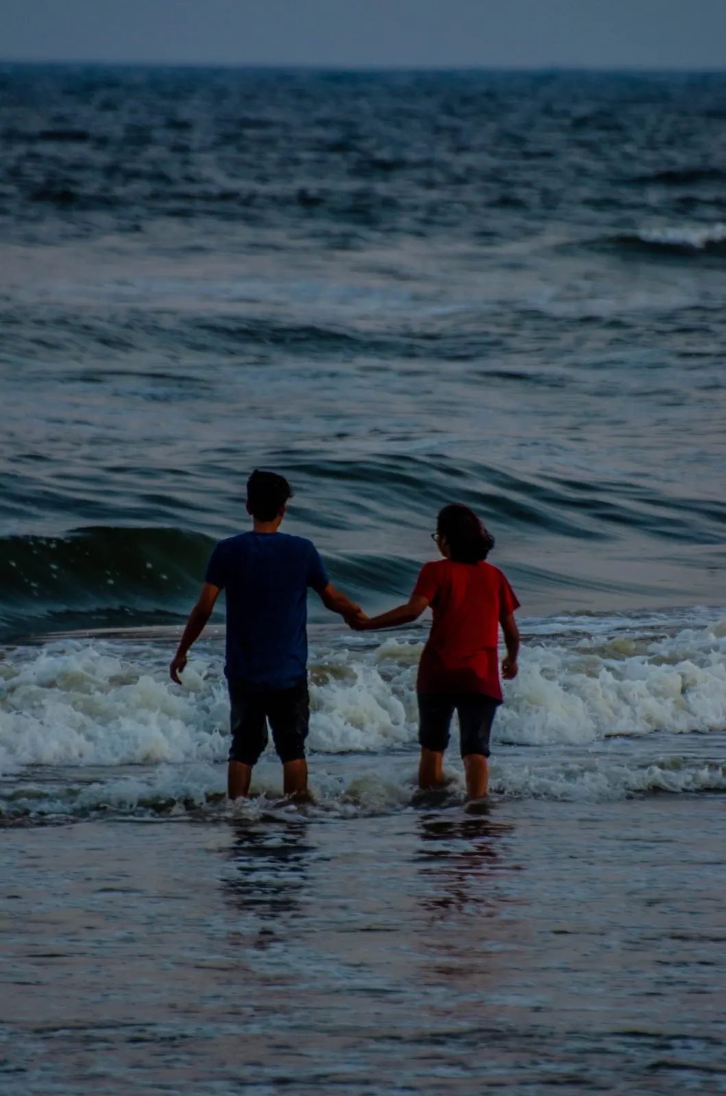 Two people holding hands walking into dark blue surf at dusk