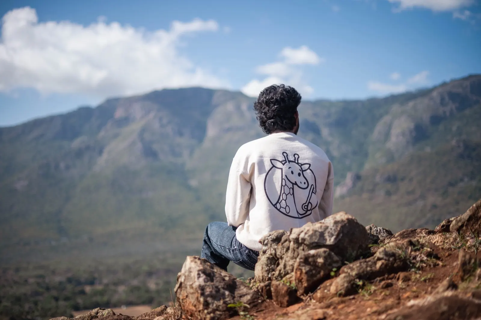 Man in a cream sweatshirt sitting on rocks, looking out at hazy green mountains under a blue sky