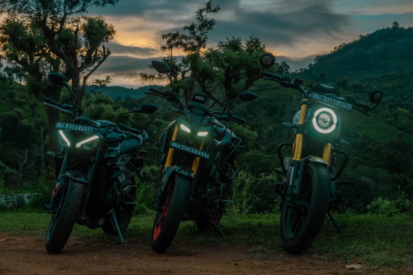 Three motorcycles with headlights glowing at blue hour against ember sunset clouds and dark hills