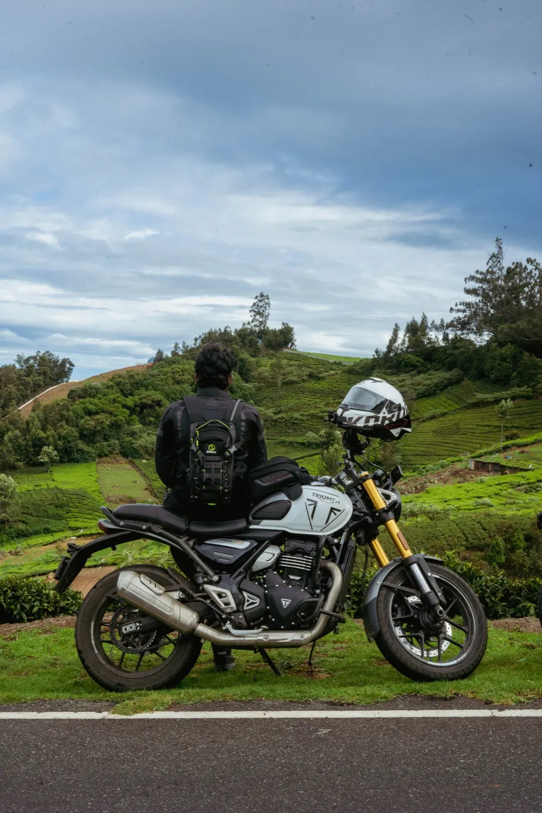 Rider seated on a parked Triumph with his back to camera, overlooking green tea-estate hills under a cloudy sky