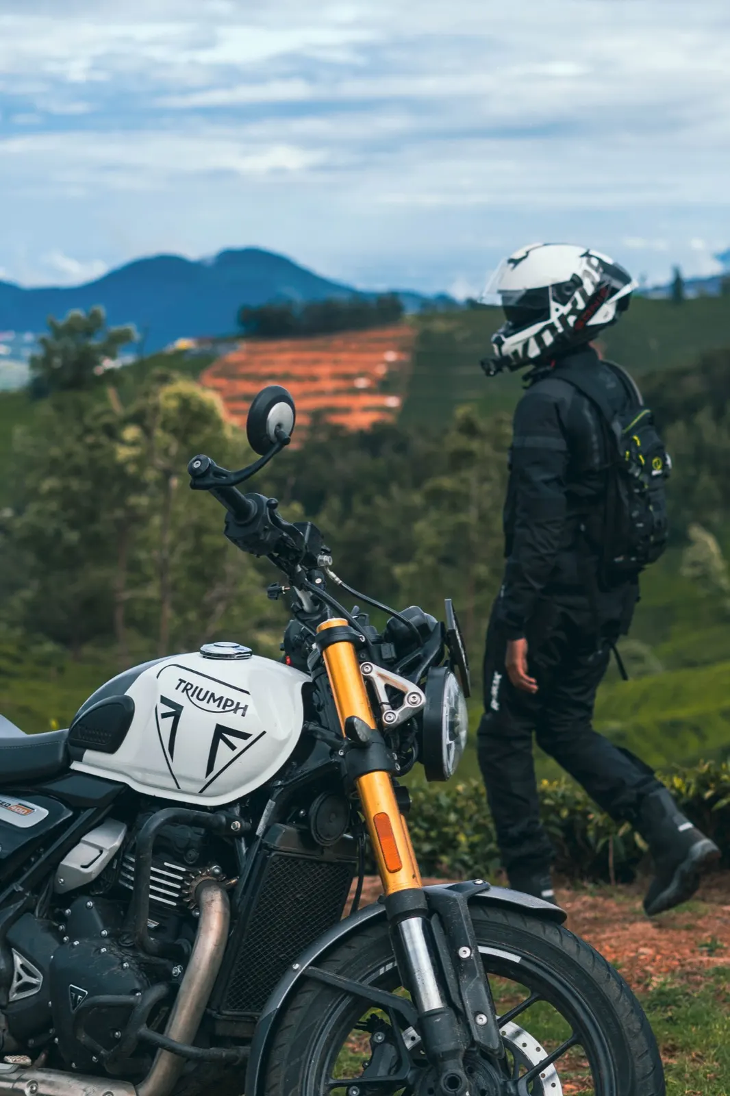 White-helmeted rider walking past a Triumph's gold forks toward terraced tea plantations and blue mountains