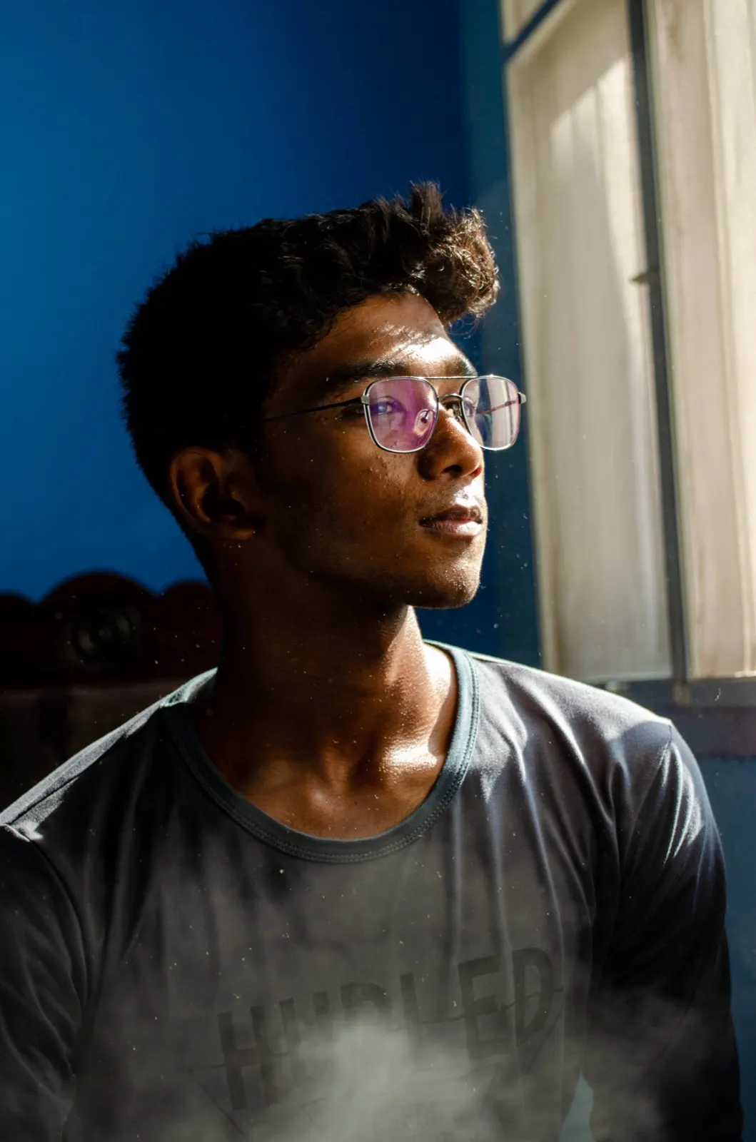Portrait of a young man in glasses lit by hard window light against a blue wall